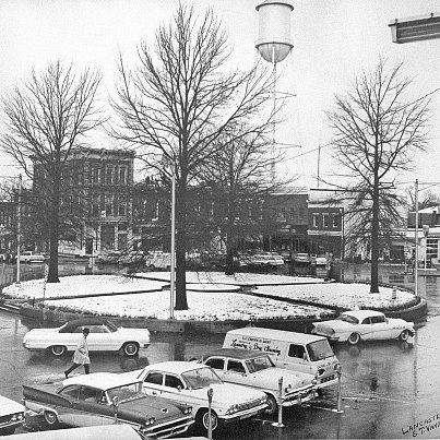 Historic black-and-white photograph of a town square in Lancaster, Kentucky, featuring classic cars parked around a circular median with leafless trees.